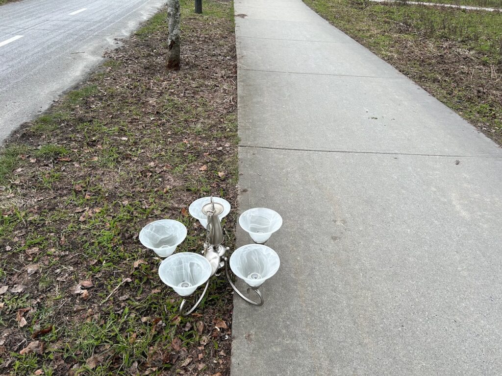 A simple chandelier with light bulbs missing, sitting strangely next to a walking path on the side of a road.