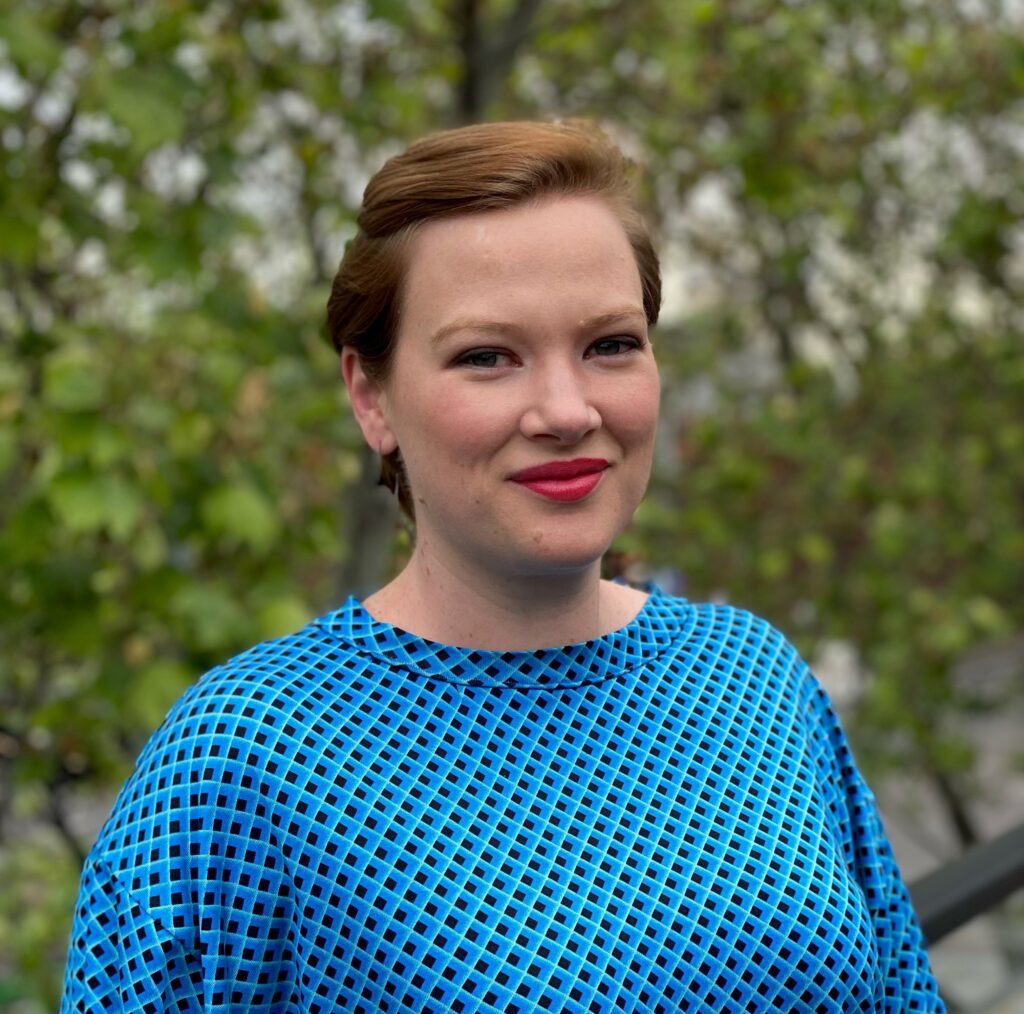 A white woman with short auburn hair, wearing red lipstick and a blue geometric print top, stands in front of a tree. She is trying very hard to look professional and not laugh at the person taking the photo.