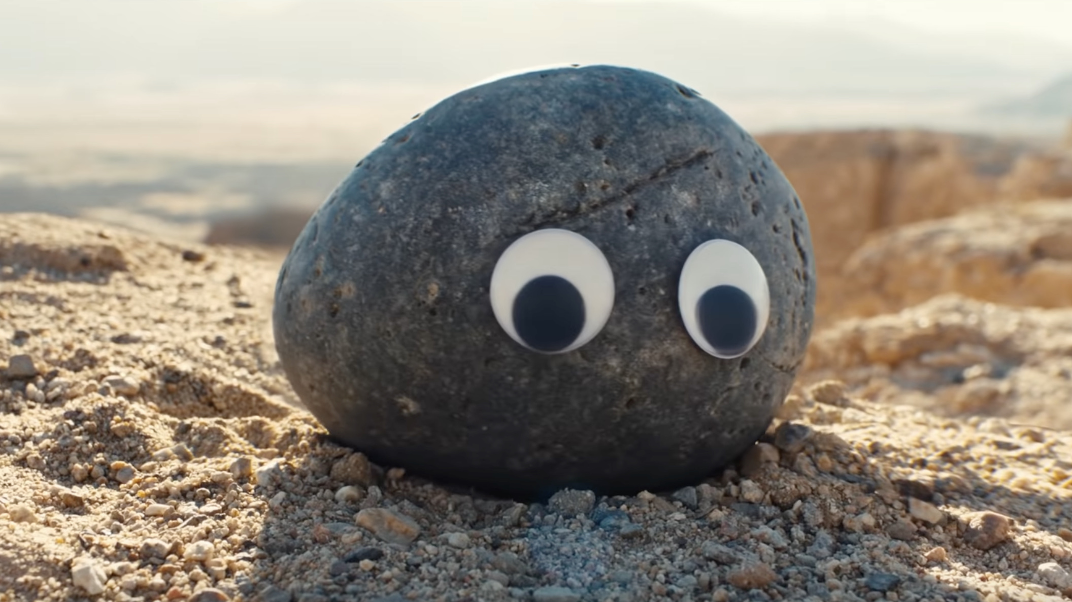 A smooth gray rock with two googly eyes sitting on a field of pebbles.