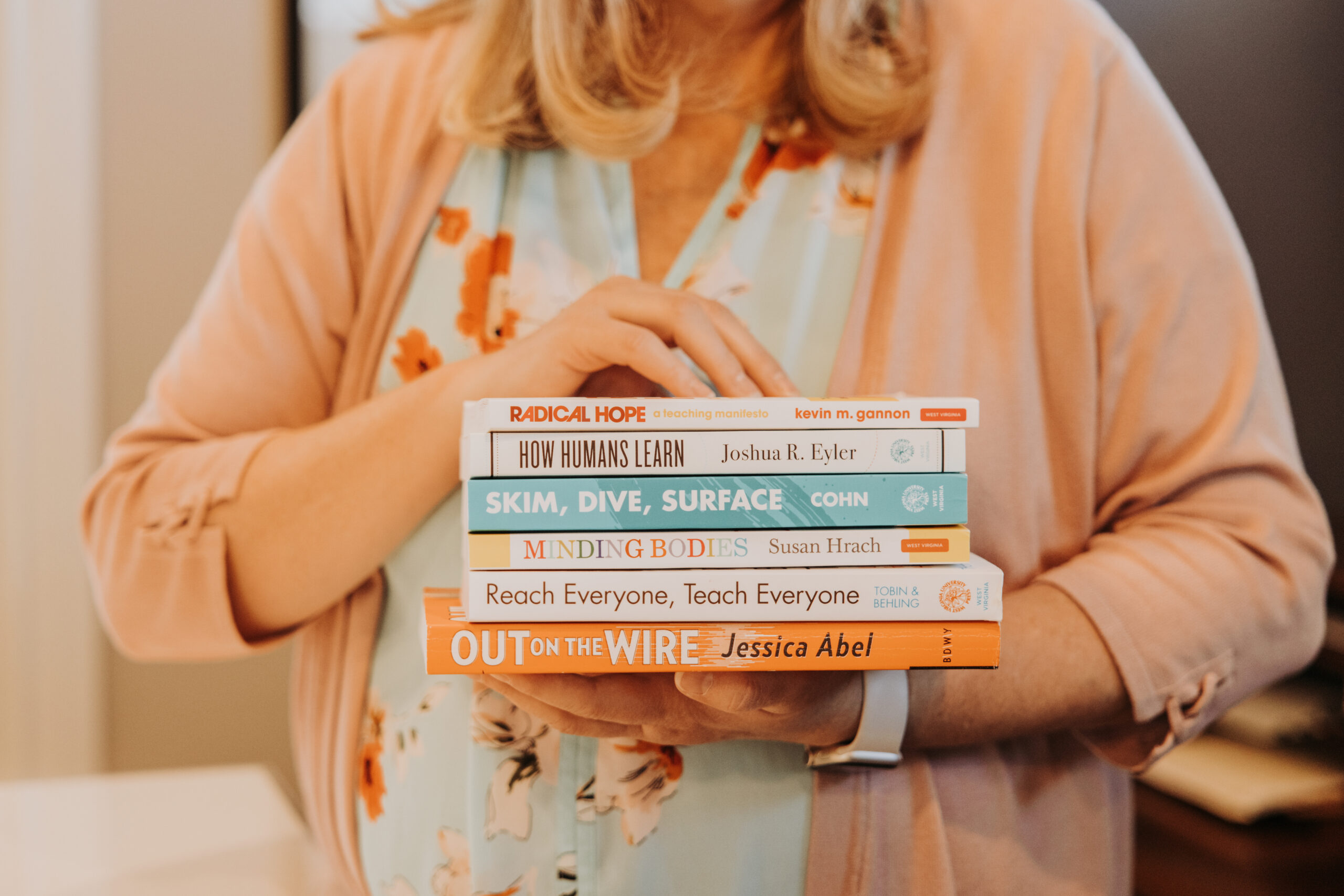 Woman holds a stack of books in front of her. Titles on the spines: Radical Hope; How Humans Learn; Skim, Dive, Surface; Minding Bodies; Reach Everyone, Teach Everyone; Out on a Wire